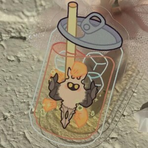 May include: A clear acrylic charm depicting a cartoon character inside a beverage cup with a straw and ice cubes. The character has a light peach body, dark brown horns, and orange accents. The cup has a silver lid.