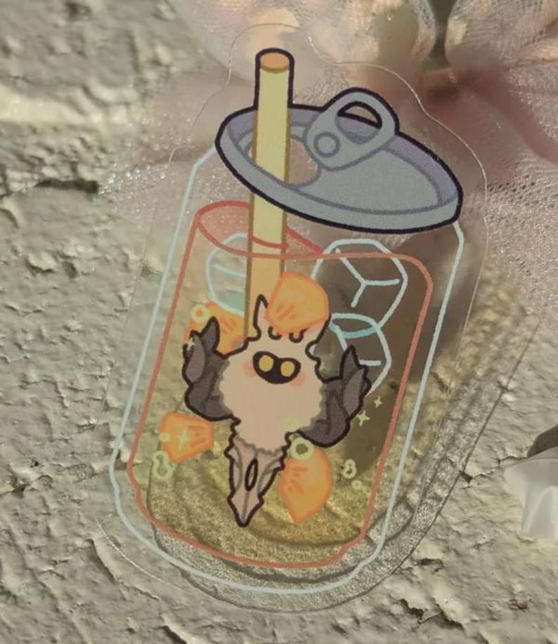 May include: A clear acrylic charm depicting a cartoon character inside a beverage cup with a straw and ice cubes. The character has a light peach body, dark brown horns, and orange accents. The cup has a silver lid.