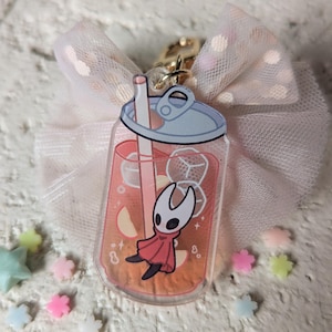 May include: A clear acrylic charm shaped like a soda can, featuring a cartoon character in a red cloak, inside a drink with ice cubes and fruit slices. The charm is attached to a gold clasp and a pink bow.