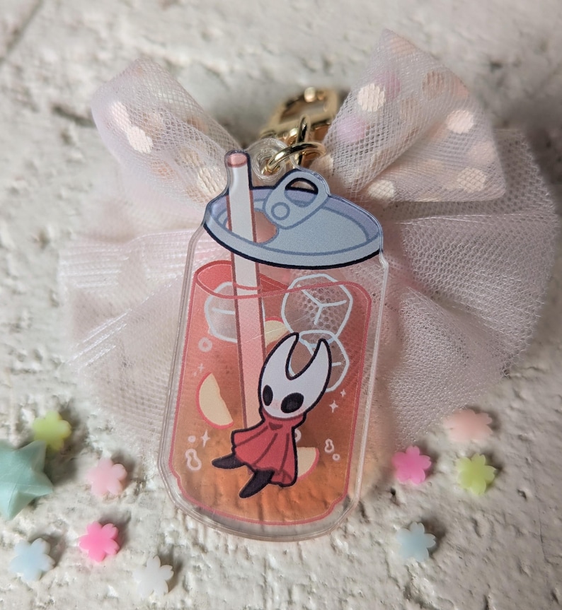 May include: A clear acrylic charm shaped like a soda can, featuring a cartoon character in a red cloak, inside a drink with ice cubes and fruit slices. The charm is attached to a gold clasp and a pink bow.
