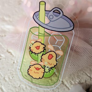 May include: A clear sticker depicting a cartoon drink in a can. The drink is green with kiwi slices and three yellow, smiling characters. The can has a silver lid and a yellow straw.