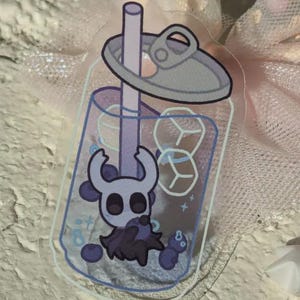 May include: A clear sticker depicting a cartoon character in a bubble tea. The drink is purple with ice cubes and a straw. The character is a white and purple creature with large eyes and horns. The drink is topped with a grey lid.