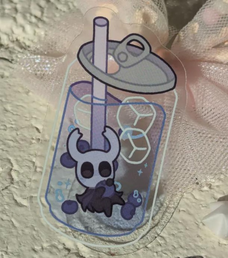 May include: A clear sticker depicting a cartoon character in a bubble tea. The drink is purple with ice cubes and a straw. The character is a white and purple creature with large eyes and horns. The drink is topped with a grey lid.