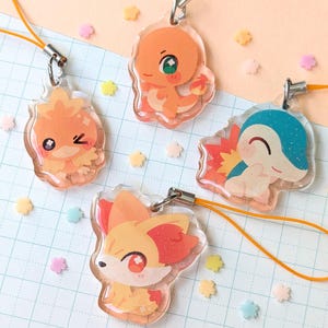 Op de afbeelding: Vier heldere acryl bedels met cartoonfiguren in oranje, geel en blauw. Elke bedel heeft een zilveren sluiting en een oranje koord. De bedels liggen op een geruite papieren achtergrond met pastelkleurige bloemvormige confetti.