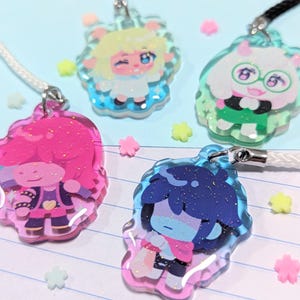 May include: Four colorful acrylic charms with cartoon character designs. Each charm is attached to a lanyard. The charms feature pink, blue, and green glitter accents. The background is a light blue surface with star-shaped decorations.