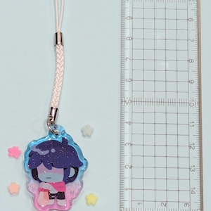 May include: A charm featuring a cartoon character with dark blue hair and a pink shirt, attached to a white cord. The charm is approximately 1.5 inches in height. The charm is surrounded by small star-shaped decorations.