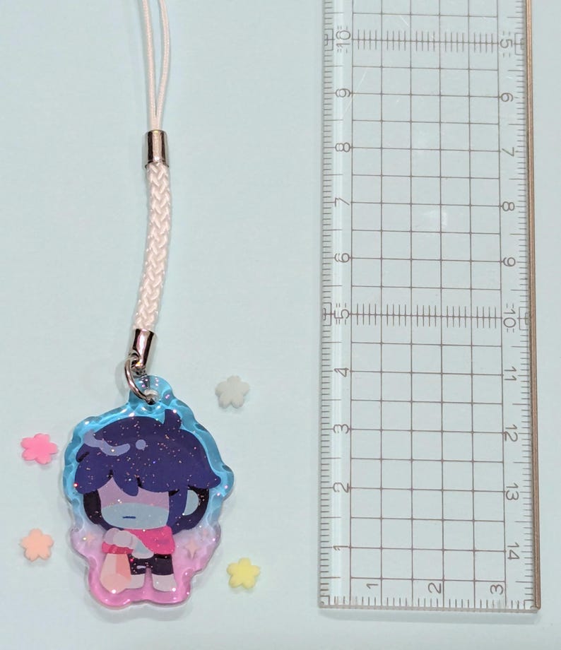 May include: A charm featuring a cartoon character with dark blue hair and a pink shirt, attached to a white cord. The charm is approximately 1.5 inches in height. The charm is surrounded by small star-shaped decorations.