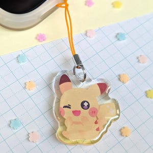 Op de afbeelding: Een gele Pikachu-bedel met rode accenten en een knipoog. De bedel is bevestigd aan een oranje koord en een zilveren sluiting. De achtergrond bevat kleurrijke bloemvormige confetti.