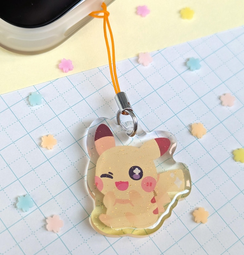 Op de afbeelding: Een gele Pikachu-bedel met rode accenten en een knipoog. De bedel is bevestigd aan een oranje koord en een zilveren sluiting. De achtergrond bevat kleurrijke bloemvormige confetti.