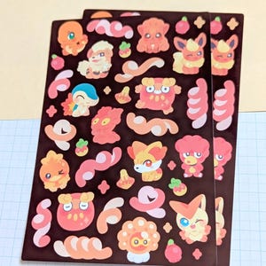 May include: A rectangular sticker sheet featuring a dark brown background with various small, colorful cartoon creatures in shades of orange, pink, and cream. The designs include multiple fire-themed characters, swirling cloud shapes, and small star icons.