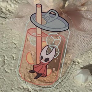 May include: A clear, rectangular sticker depicting a cartoon character in a drink. The peach-coloured beverage contains ice cubes and fruit slices. The character wears a red cape and a white mask.