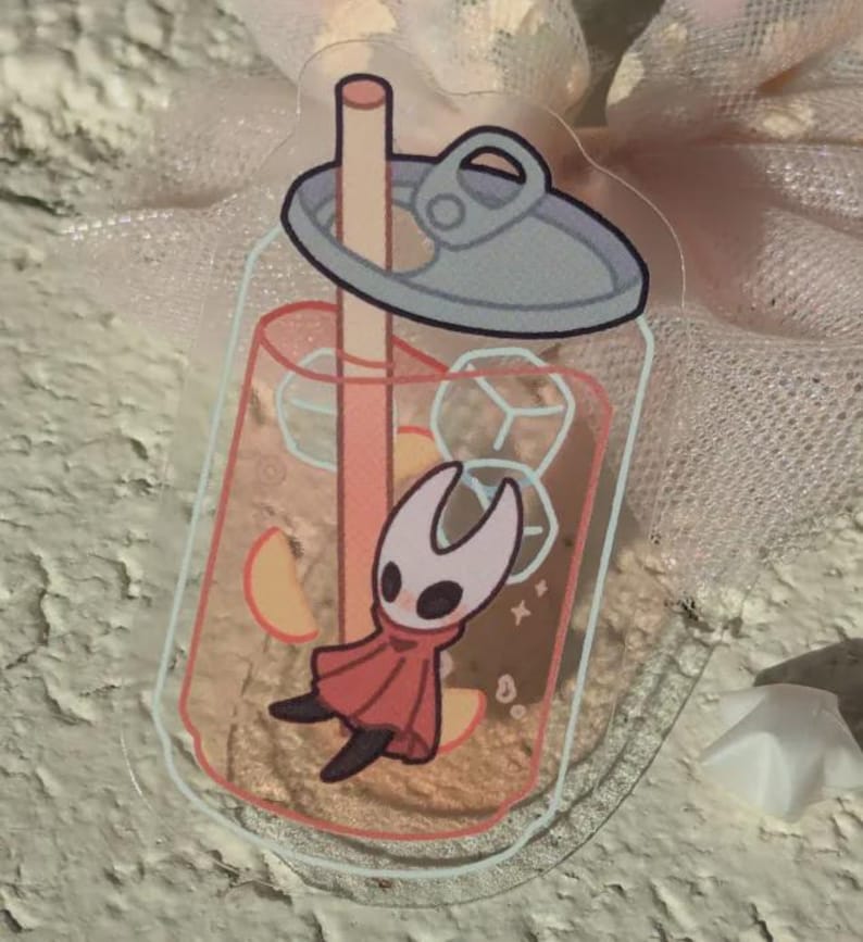 May include: A clear, rectangular sticker depicting a cartoon character in a drink. The peach-coloured beverage contains ice cubes and fruit slices. The character wears a red cape and a white mask.