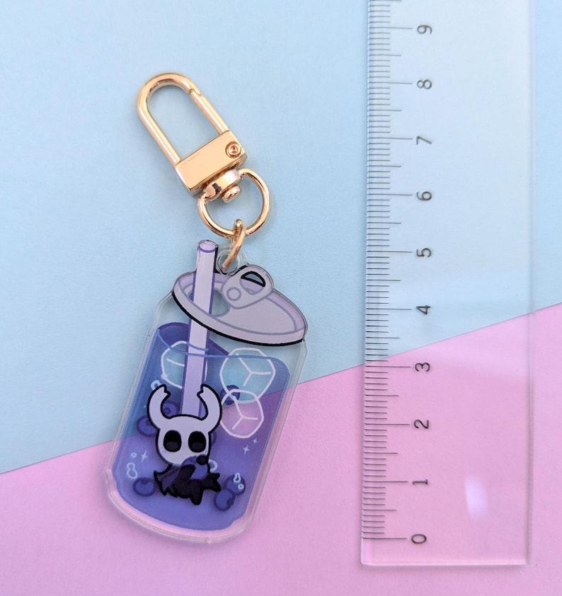 May include: A clear acrylic keychain shaped like a bubble tea cup, featuring a cartoon character and ice cubes in a purple liquid. The keychain has a gold-colored clasp and measures approximately 3 inches in height.
