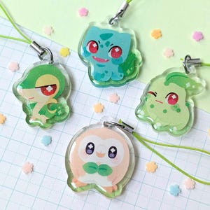 Op de afbeelding: Vier heldere acryl bedels met kleurrijke cartoonfiguren. De bedels zijn bevestigd aan groene koorden met zilverkleurige sluitingen. De figuren zijn in tinten groen, blauw en perzik, met glitteraccenten. De achtergrond is een lichtgroen en wit raster.