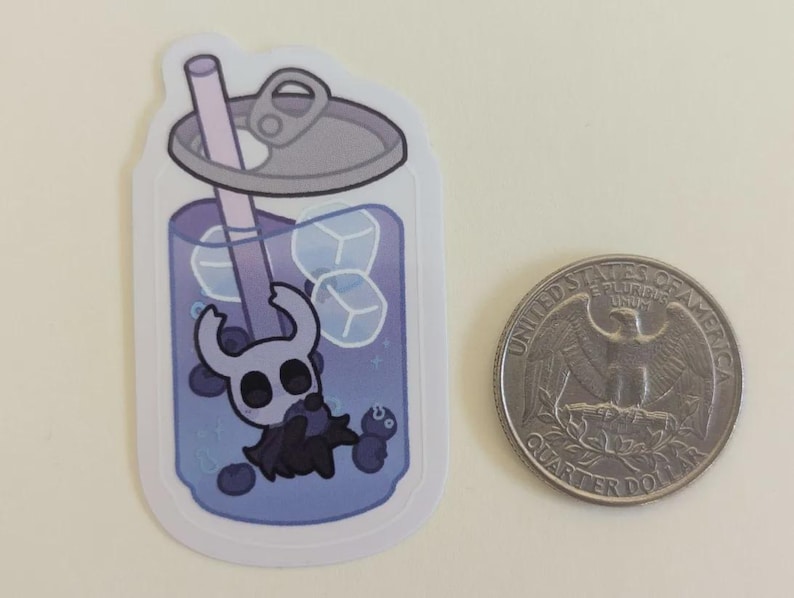 May include: A sticker depicting a cartoon character in a blue drink can with ice cubes and a straw. A quarter dollar coin is next to the sticker.