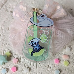 May include: A clear acrylic charm shaped like a soda can, featuring a cartoon character inside. The charm has a gold-colored clasp and is set against a pink tulle bow and scattered pastel-colored star and flower-shaped decorations.