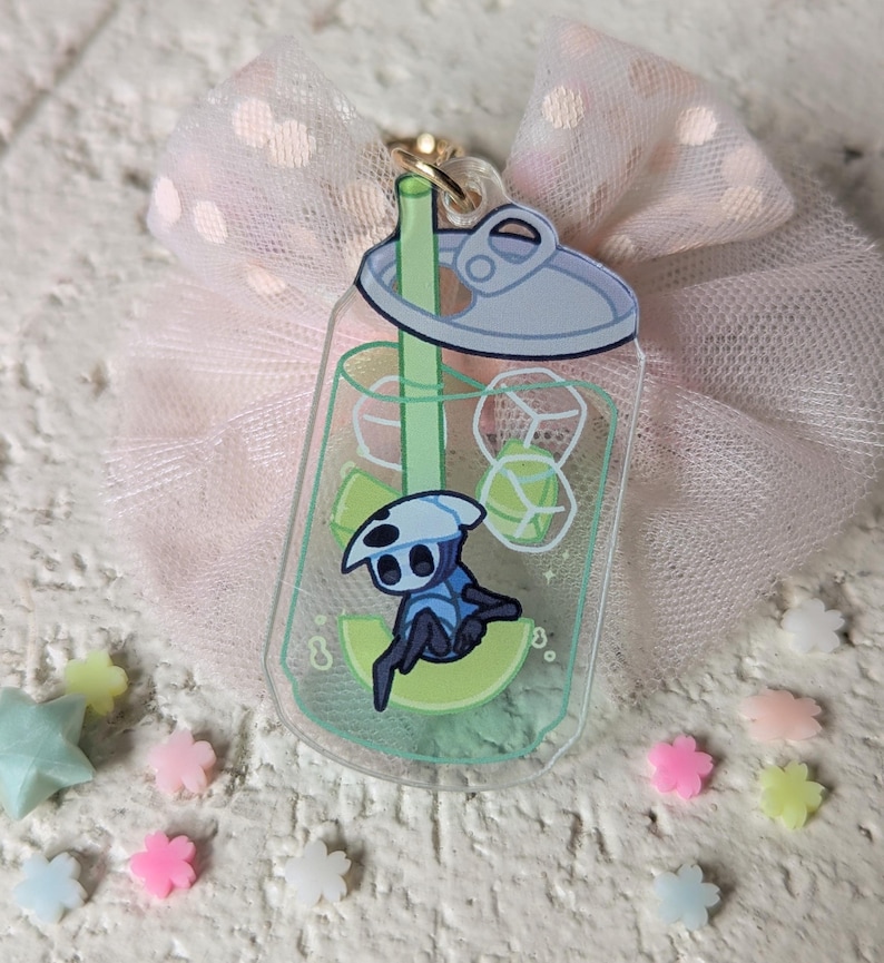 May include: A clear acrylic charm shaped like a soda can, featuring a cartoon character inside. The charm has a gold-colored clasp and is set against a pink tulle bow and scattered pastel-colored star and flower-shaped decorations.