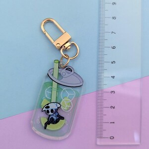 May include: A clear acrylic keychain featuring a cartoon character in a soda cup design. The keychain has a gold-colored clasp and ring. The soda cup design includes ice cubes, a straw, and a lime slice. The ruler next to the keychain shows it is approximately 3 inches long.