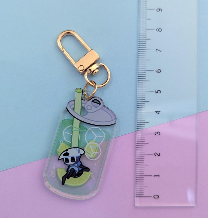 May include: A clear acrylic keychain featuring a cartoon character in a soda cup design. The keychain has a gold-colored clasp and ring. The soda cup design includes ice cubes, a straw, and a lime slice. The ruler next to the keychain shows it is approximately 3 inches long.