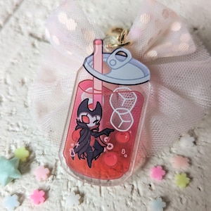 May include: A clear acrylic charm in the shape of a bubble tea drink, featuring a cartoon character in a black cape. The drink is red with ice cubes and a straw. A pink bow and small star and flower-shaped decorations surround the charm.