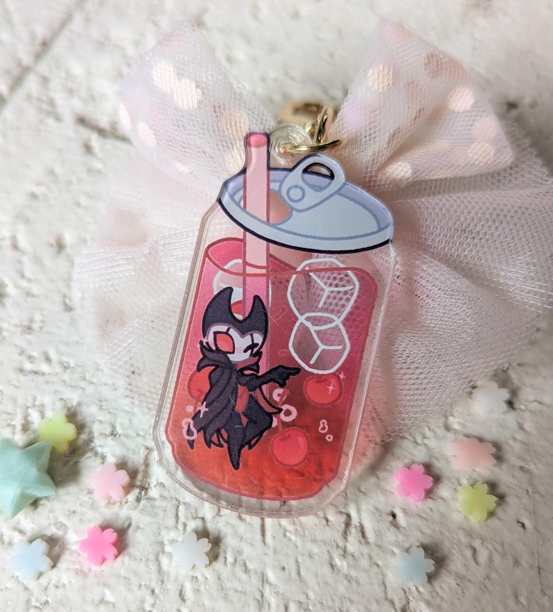 May include: A clear acrylic charm in the shape of a bubble tea drink, featuring a cartoon character in a black cape. The drink is red with ice cubes and a straw. A pink bow and small star and flower-shaped decorations surround the charm.