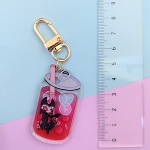 May include: A clear acrylic keychain featuring a cartoon character inside a red drink with ice cubes and a straw. The keychain has a gold-colored clasp. The ruler next to the keychain measures up to 9 inches.