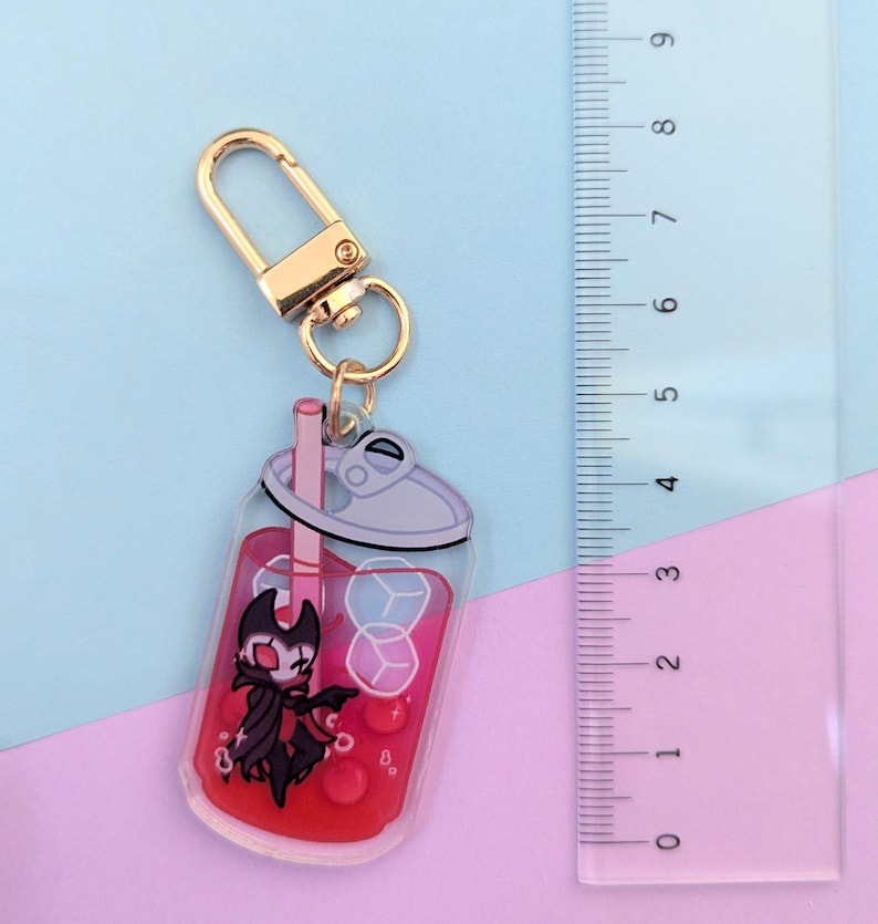 May include: A clear acrylic keychain featuring a cartoon character inside a red drink with ice cubes and a straw. The keychain has a gold-colored clasp. The ruler next to the keychain measures up to 9 inches.