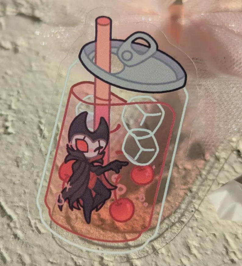 May include: A clear sticker with a cartoon character inside a soda can design. The character is dark with red accents, holding cherries. The can has a pink straw and ice cubes. The design is outlined in white.