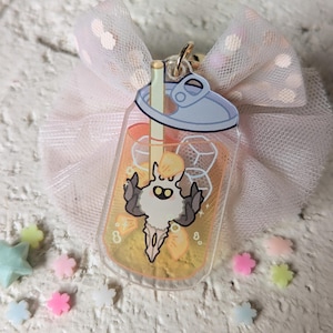 May include: A clear acrylic charm in the shape of a beverage can, featuring a cartoon character inside. The charm has a gold-colored clasp and is attached to a pink bow with white polka dots. Scattered around are small, colorful star and flower-shaped decorations.