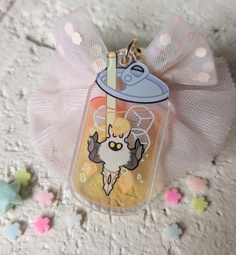 May include: A clear acrylic charm in the shape of a beverage can, featuring a cartoon character inside. The charm has a gold-colored clasp and is attached to a pink bow with white polka dots. Scattered around are small, colorful star and flower-shaped decorations.