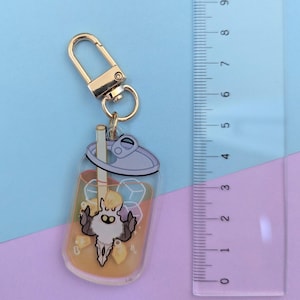 May include: A clear acrylic keychain featuring a cartoon beverage with ice cubes and a straw. The keychain has a gold-colored clasp and a small, stylized character in the center. The drink is orange-colored.