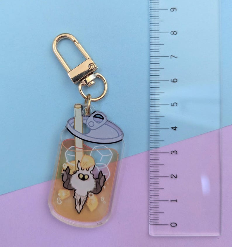 May include: A clear acrylic keychain featuring a cartoon beverage with ice cubes and a straw. The keychain has a gold-colored clasp and a small, stylized character in the center. The drink is orange-colored.