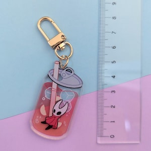 May include: A clear acrylic keychain featuring a red drink with ice cubes and a cartoon character. The keychain has a gold-colored clasp and a metal ring. The ruler next to the keychain measures up to 9 inches.