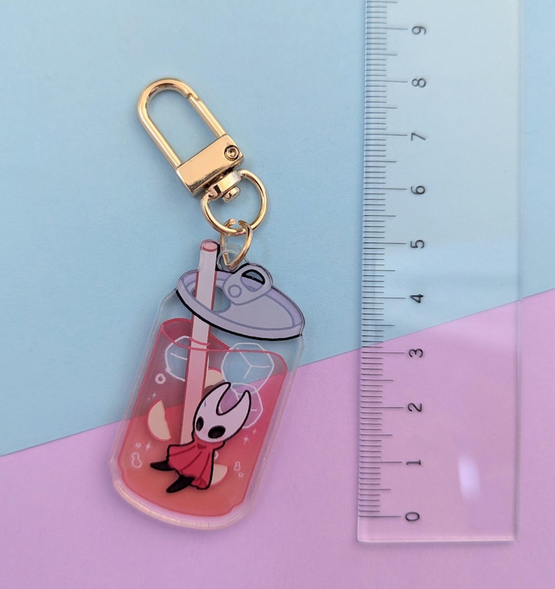 May include: A clear acrylic keychain featuring a red drink with ice cubes and a cartoon character. The keychain has a gold-colored clasp and a metal ring. The ruler next to the keychain measures up to 9 inches.