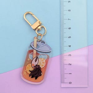 May include: A clear acrylic keychain featuring a cartoon drink with orange slices and ice cubes. The keychain has a gold-colored clasp and a cartoon character with a black cape. The ruler next to the keychain measures up to 9 inches.