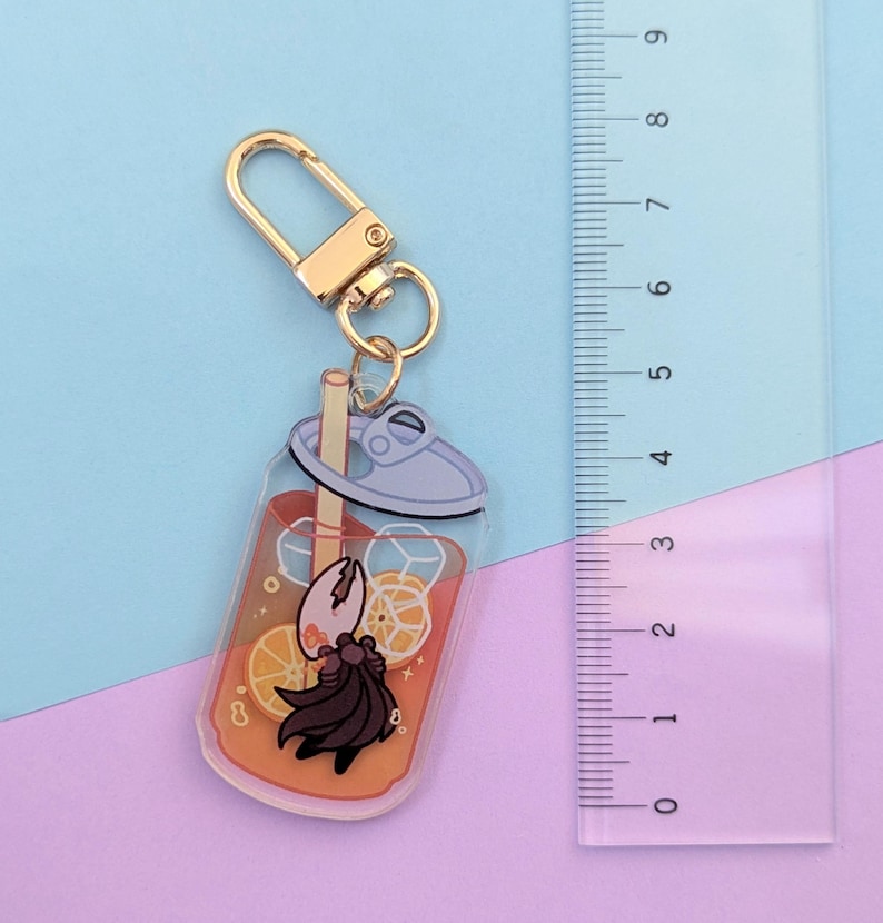 May include: A clear acrylic keychain featuring a cartoon drink with orange slices and ice cubes. The keychain has a gold-colored clasp and a cartoon character with a black cape. The ruler next to the keychain measures up to 9 inches.