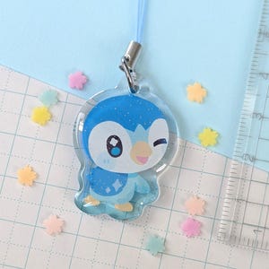 Op de afbeelding: Een heldere acryl bedel met een blauw-witte cartoonpingu&iuml;n die knipoogt. De bedel heeft een blauw koord en een zilveren sluiting. Er liggen kleurrijke stervormige confetti verspreid. De achtergrond is lichtblauw.