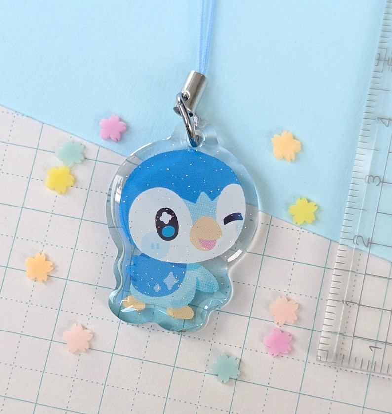 Op de afbeelding: Een heldere acryl bedel met een blauw-witte cartoonpingu&iuml;n die knipoogt. De bedel heeft een blauw koord en een zilveren sluiting. Er liggen kleurrijke stervormige confetti verspreid. De achtergrond is lichtblauw.