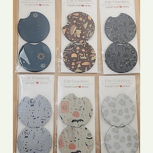 Op de afbeelding: Zes sets met auto-onderzetters met verschillende Halloween-thema's. Elk set bevat twee ronde onderzetters met een witte achtergrond en zwarte contouren. De ontwerpen omvatten paddenstoelen, katten, sterren, pompoenen, vleermuizen en geesten. De tekst 'Car Coasters' en 'Handmade with love' staat op elk set gedrukt.