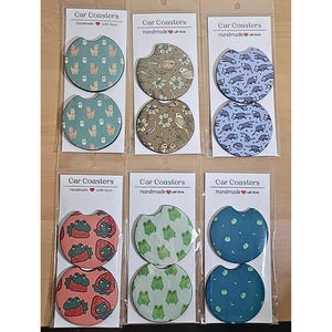 May include: Six sets of car coasters with various designs. Each set includes two round coasters with a different design. The designs include owls, birds, raccoons, strawberries, and frogs. The coasters are handmade with love.