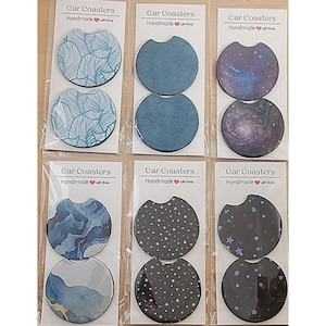 May include: Six sets of car coasters, each set containing two coasters. The coasters are round and feature different designs, including blue and white waves, a galaxy, and a black background with gold stars. The packaging is clear plastic and each set has a label that reads "Car Coasters Handmade with love".
