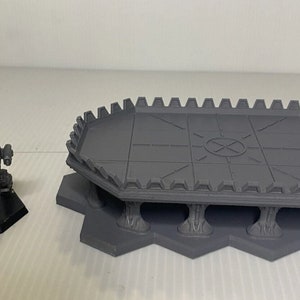 May include: A gray miniature battle mech figure on a black hexagonal base next to a gray hexagonal platform with a detailed surface and crenelated edge. The platform is supported by pillars.