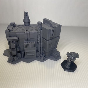 以下が含まれることがあります： パイプや塔などの詳細な建築要素を備えた、グレーの3Dプリントされた未来的な建物のミニチュア。小さなロボットフィギュアが黒い六角形の台座に立っています。白い表面に配置されています。