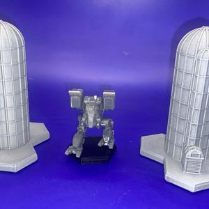 May include: A gray, detailed model of a mech robot standing on a black base. Two gray, detailed models of silos with bases are on either side of the mech.