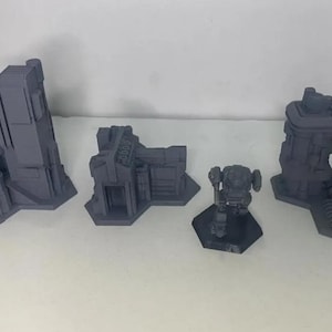 Puede incluir: Cinco edificios en miniatura impresos en 3D y una figura de robot, todos de color gris oscuro. Los edificios son de varias formas y tamaños, algunos tienen ventanas y puertas. La figura del robot está de pie sobre una base hexagonal.