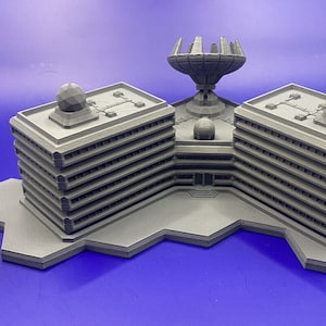 Puede incluir: Modelo gris impreso en 3D de un complejo urbano futurista, con dos edificios de varios pisos conectados por una estructura central con una antena parabólica. Una pequeña figura de robot está cerca. El modelo está sobre una superficie azul.