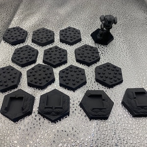 Puede incluir: Una colección de bases hexagonales negras en miniatura con superficies texturizadas y una pequeña figura oscura sobre una base a juego. Las bases están dispuestas sobre una superficie plateada reflectante, lo que sugiere un tema de juego de mesa o modelismo.