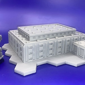 May include: A grey, 3D-printed model of a building with a flat roof and multiple windows. Beside it is a grey robot model on a black base. The models are on a light grey platform against a blue background.