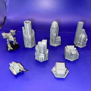 May include: A collection of gray 3D printed miniature buildings and vehicles, including a tank, a mech, and several skyscrapers. The buildings are all hexagonal in shape and have a futuristic design.