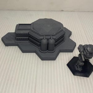 May include: A gray 3D printed model of a futuristic building with a hexagonal base and a gray 3D printed model of a robot on a hexagonal base.